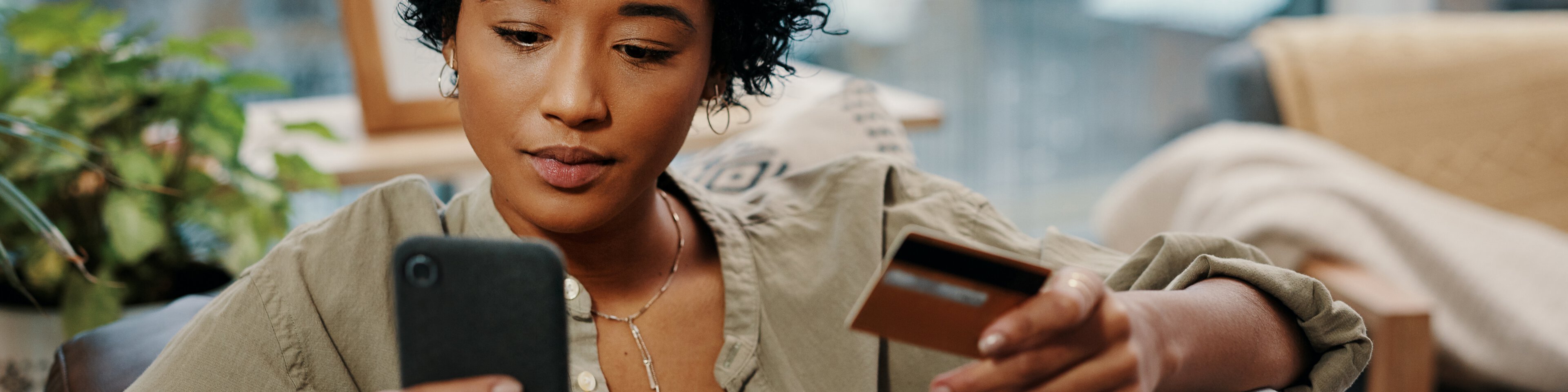 Eine junge Frau hält ihre Kreditkarte in der Hand und schaut auf ihr Handy | © Delmaine Donson - YURI ARCURS PRODUCTIONS